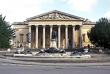 A Palladian style nineteenth century stone building with a large colonnaded porch. In front a large metal statue on a pedestal and fountains with decorations.