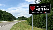 A large rectangular metal sign, mostly black, with the words "Welcome To Virginia" and "Virginia is for lovers" with a red heart symbol on the left.