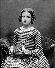 Three quarter length studio photo of seated girl about nine years old, looking slightly plump and rather solemn, in a striped dress, holding a basket of flowers on her lap.
