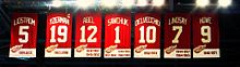 The banners of seven retired numbers. The banners, from left to right, read "Lidström 5" "Yzerman 19" "Sawchuk 1" "Delvecchio 10" "Lindsay 7" "Abel 12" "Howe 9". The Yzerman banner has a small C at the top right corner.