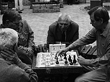 Photo shows two men playing chess while two more look on.