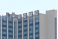 Looking up at a hospital with blue and teal window panes, four square signs read "E C M C" in white with a light gray background