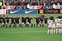 Rugby team wearing all black, facing the camera, knees bent, and facing toward a team wearing white