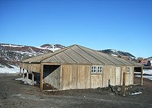 Wooden structure with door and two small windows. To the left is an open lean-to. In the background are partly snow-covered mountains.