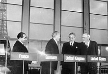 Four men stand behind podiums with their country names of France, Germany, United Kingdom, and United States, in front of a backdrop of the Eiffel Tower.