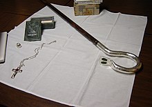 A crosier, pectoral cross, episcopal ring, and jade box on top of a white cloth that covers a dark brown wooden table