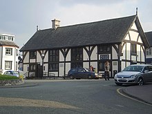 A half timbered building of two floors, with four sets of leaded windows to the front aspect and one set to the side. The build has a steep, slate roof, with a single chimney placed left of centre. Steps and a ramp lead up to its single visible entrance