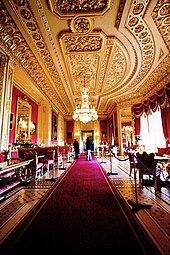 A photograph of a large room with a long red carpet stretching through the middle of it and windows on the right hand side. Furniture fills both sides of the room. The ceiling contains ornate plasterwork and a chandelier hangs down from the middle of the picture.