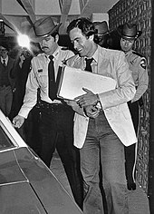 A smiling Bundy holds a sheaf of papers and enters a vehicle. He is escorted by two police officers.