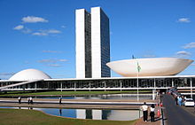 The view shows a 20th-century building with two identical towers very close to each other rising from a low building which has a dome at one end, and an inverted dome, like a saucer, at the other.