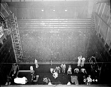 Tall square industrial room seen from above. Its cement walls have metal ladders and meshes, and a dozen people work on the floor.