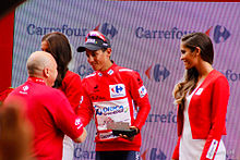 A photograph of Esteban Chaves, wearing the race leader's red jersey, shaking hands with an official, with podium girls either side