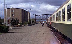 View of a train stopped at a long railway platform, at the end of which is an arched iron bridge. A grey concrete barracks and East German state emblem are visible on the side of the platform. Several people are standing or walking on the platform and the train's doors stand open.