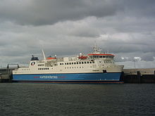 A large modern ferry with a blue hull and white topsides lies next to a harbour under grey skies.