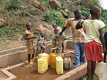 Photograph depicting one adult and five children filling jerrycans at a rural metal water pump with concrete base, at the bottom of a steep rocky hillside