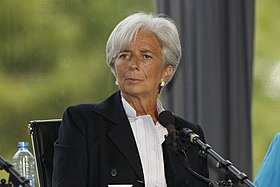 A three-quarter portrait of an elegantly dressed Christine Lagarde, perhaps in her early 60s sitting in a chair behind a microphone. She looks fit and tanned. Her overall mien is alert, pleasant, and intelligent.