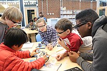 Five middle school students work together at a table using a soldering iron.