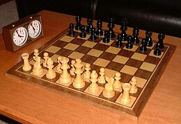 This photo shows a chessboard with pieces set up on both sides, ready to play. A chess clock is at the side.