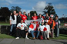 A group of thirteen supporters pose together, nine standing in back row, four seated at front, some wearing rugby jerseys and others sporting traditional Japanese costumes and Japanese flags.