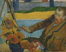 A seated red-bearded man wearing a brown coat, facing to the left, with a paintbrush in his right hand, is painting a picture of large sunflowers.