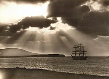 A sepia-tone photograph of a 3-masted 19th-century sailing vessel at sea with the shore in the foreground and dark hills beyond. The sun's rays cast light and shadows in the sky as they break through the clouds.