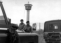 Two British soldiers carrying rifles standing behind a pair of Land-Rover vehicles, one of which has a "British Frontier Service" plate. Behind them is a high mesh fence, behind which is a tall watchtower with an octagonal cabin at its top.