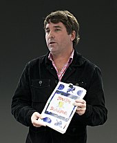 Photograph of Stephen Hillenburg standing holding a book with the title SpongeBob SquarePants looking to his right