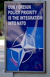 A blue poster at a bus stop with the NATO logo and the words "OUR FOREIGN POLICY PRIORITY IS THE INTEGRATION INTO NATO" in white.