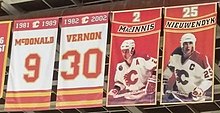 Four rectangular banners are hanging from the rafters in a hockey arena. The two on the far left are predominantly white with red and gold trim. The left one of these says "1981–1989 McDONALD 9" and the right "1982–2002 VERNON 30". On the right side of them, two more banners are shown. Both are mainly red, each showing (from top to bottom) a number, then a red banner with gold trim showing a name, and lastly a person in full hockey gear, who is shown wearing a white jersey with red trim and a white helmet. The left one of these says "2 MacINNIS", while the right one says "25 NIEUWENDYK".